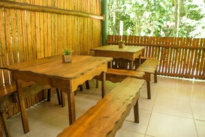 Un área de comedor al aire libre con bancos y mesas, rodeada por una valla de bambú y follaje.