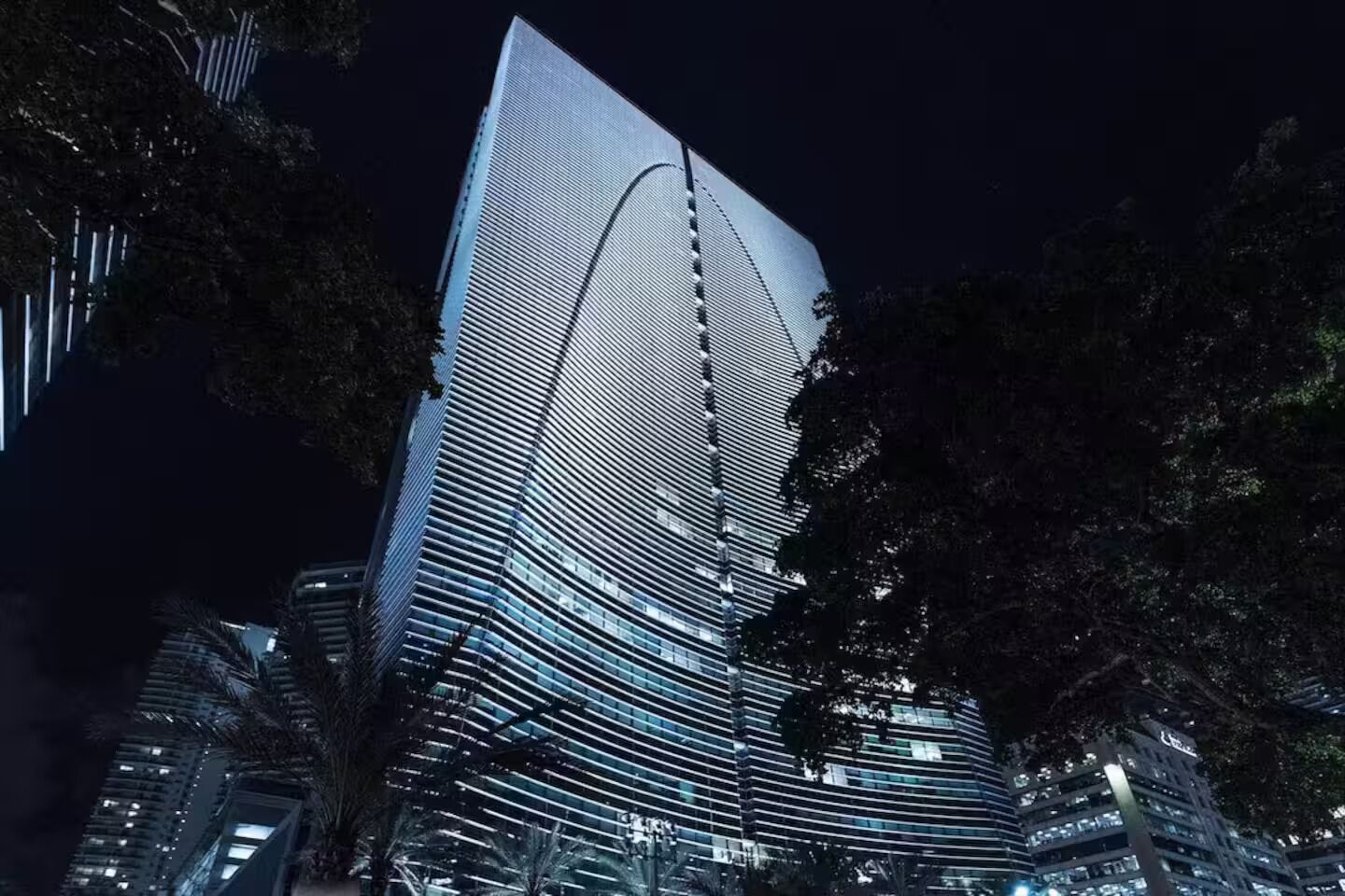A modern high-rise building with a distinctive curved glass facade, surrounded by trees.