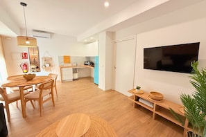 Eine moderne Küche mit Holzausbau, ein Essbereich mit rundem Tisch und Stühlen, ein an der Wand befestigter Fernseher und eine Pflanze rechts.