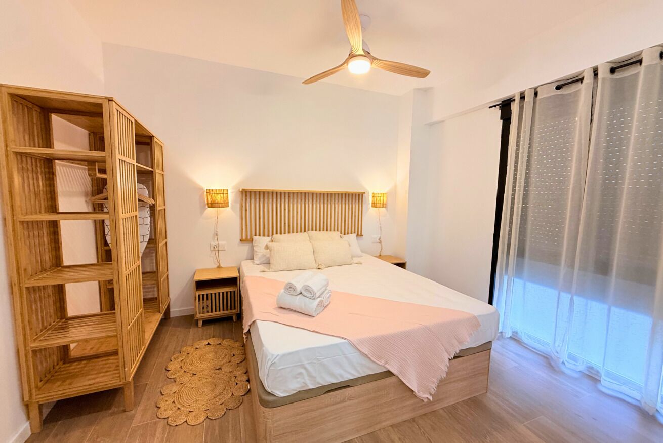 A bedroom with a bed, bedside tables, a wooden wardrobe, a ceiling fan, and a window with curtains.