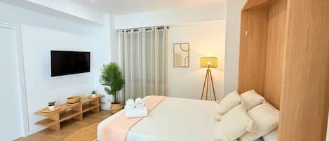 A modern hotel room with a bed, a TV, a desk with a lamp, and a potted plant.
