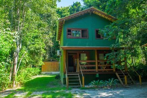 Uma casa verde com deck de madeira, rodeada por vegetação densa.