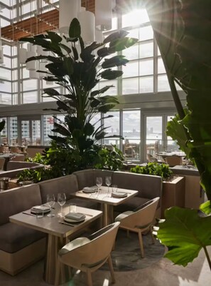 Ein modernes Restaurant mit hohen Decken, großen Fenstern und Zimmerpflanzen.