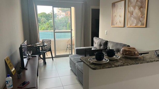 Ein modernes Wohnzimmer mit einem grauen Sofa, einem Essbereich mit Tisch und Stühlen, einer Küchenzeile mit Kuchen und Blick auf einen Pool durch eine Glastür.