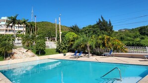 Una piscina con agua azul cristalina, rodeada por una cerca blanca, y una colina con vegetación al fondo.