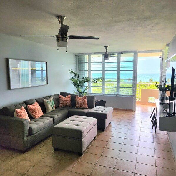 A living room with a sectional sofa, a ceiling fan, a large window with a view of the ocean, and a tiled floor. - SUNRISE IN VILLAS DEL FARO (Emajagua)