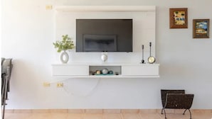 Una sala de estar con un televisor montado en la pared, un pequeño estante con adornos, un reloj y un banco.