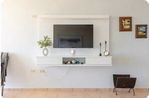 A living room with a mounted TV, a small shelf with decorative items, a clock, and a bench. - SUNRISE IN VILLAS DEL FARO (Emajagua)