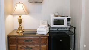 Una habitación de hotel con un aparador de madera, una lámpara, una pila de toallas, un microondas y un electrodoméstico negro, posiblemente un lavavajillas.