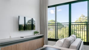 A modern living room with a flat-screen TV, a sofa, a coffee table, and a large window with a view of greenery.