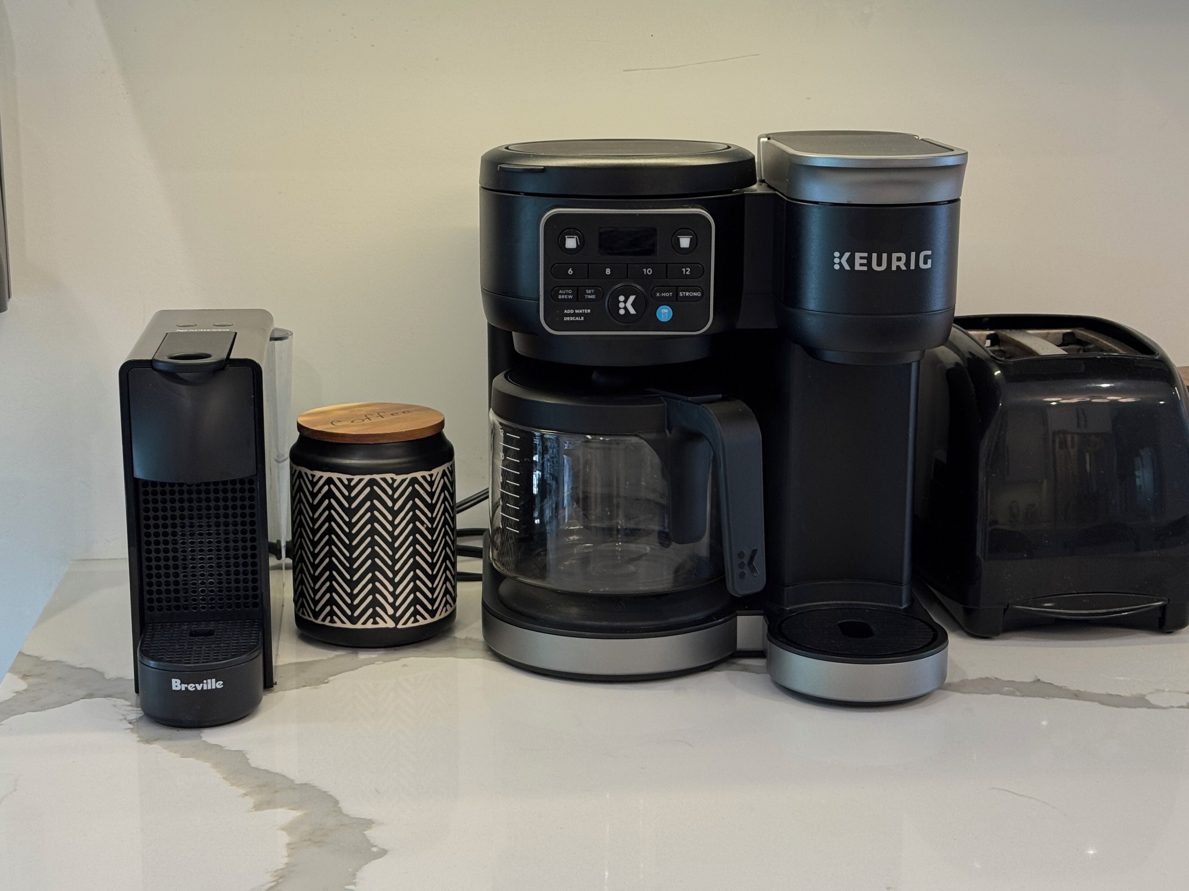 A coffee maker, a Breville coffee machine, a Keurig coffee maker, a coffee mug, and a toaster on a kitchen counter.