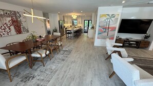 A modern living room with a dining area, a kitchen in the background, and a large TV on the right.