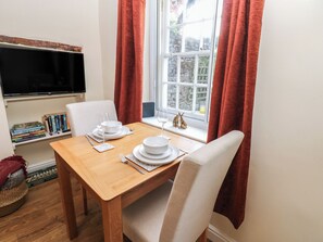 Una mesa de comedor dispuesta con platos, vasos y cubiertos, una estantería con libros, un televisor y una ventana con cortinas.