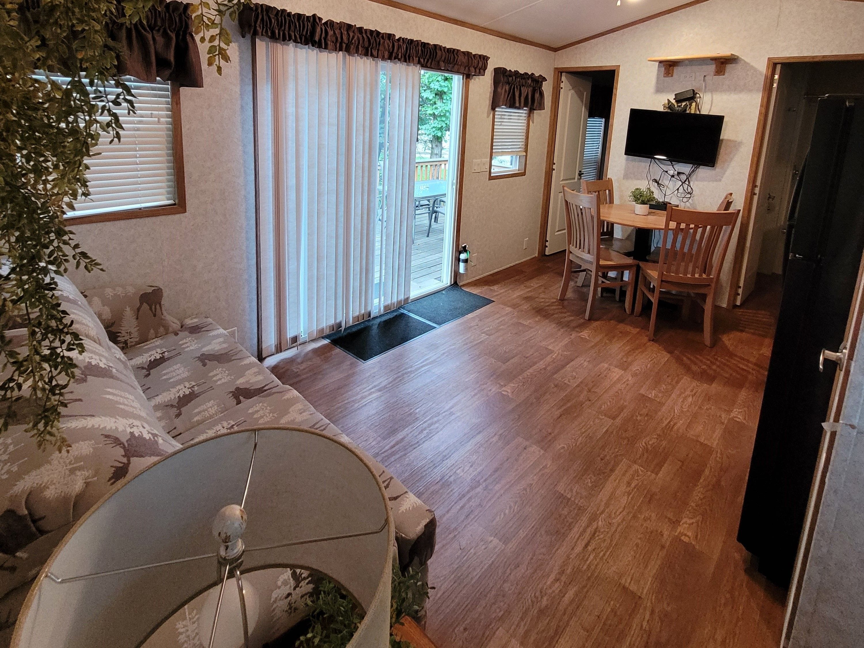 A living room with a sofa, a dining table with chairs, a television mounted on the wall, and a door leading to another room.