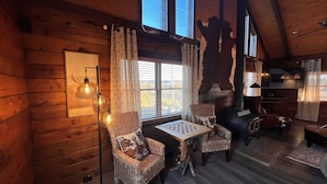 A cozy cabin interior with wooden walls, a fireplace, a dining table, and two wicker chairs. - Natural State Cabins - 5 Cabin Listing (Hot Springs)