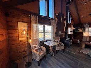A cozy cabin interior with wooden walls, a fireplace, a dining table, and two wicker chairs. - Natural State Cabins - 5 Cabin Listing (Hot Springs)