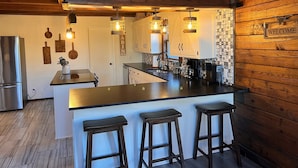 A kitchen with wooden walls, a welcome sign, and a bar area with stools. - Natural State Cabins - 5 Cabin Listing (Hot Springs)