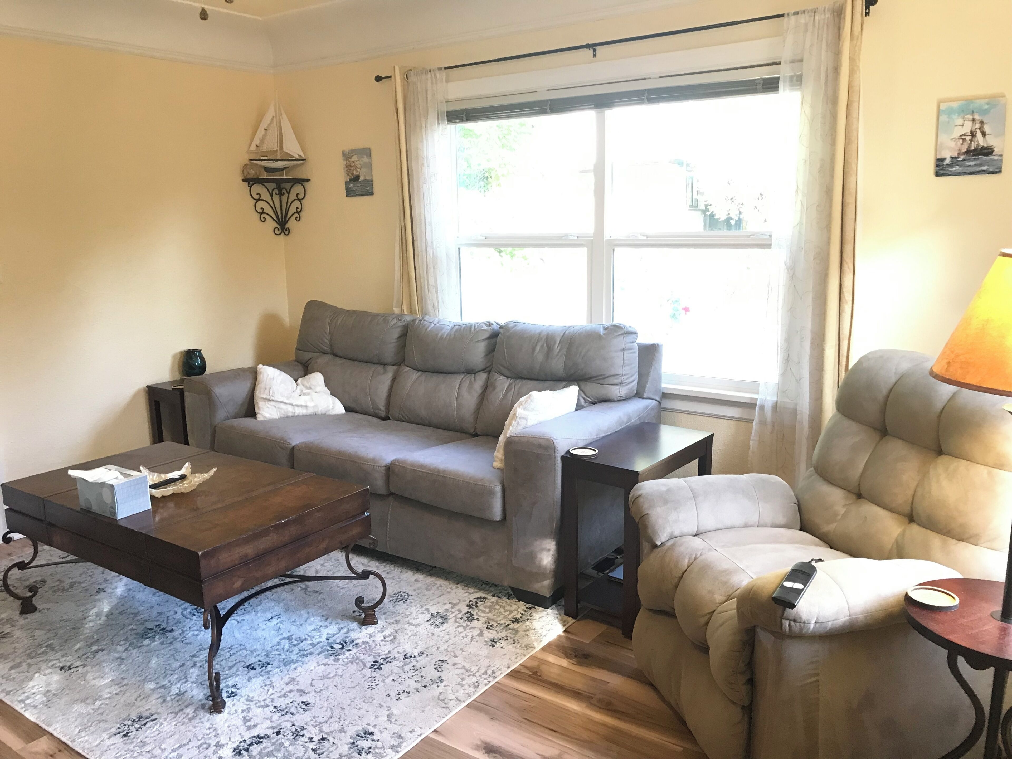 Un salon avec un canapé gris, une table basse en bois, un fauteuil beige et une fenêtre avec des rideaux.
