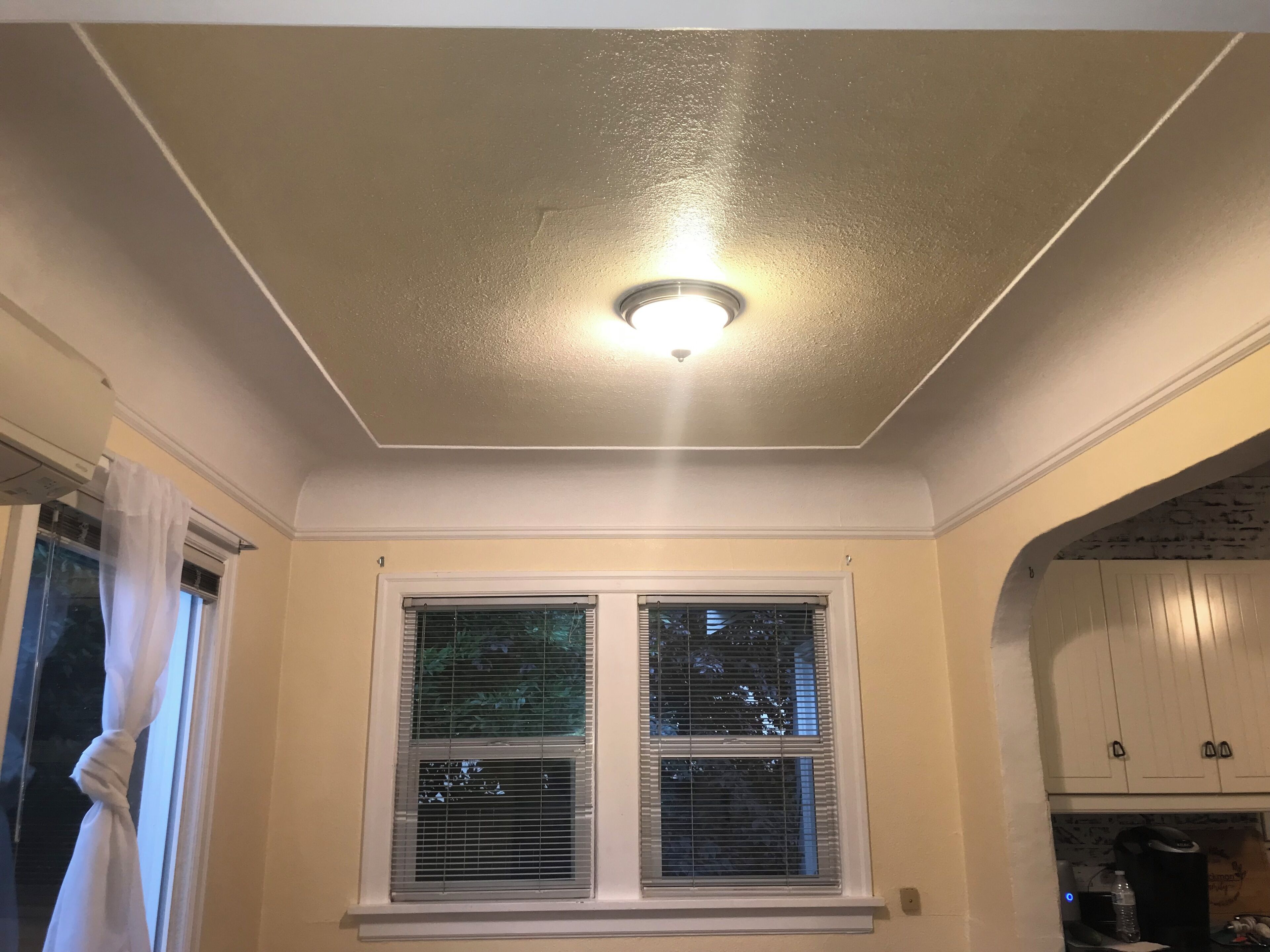 Een kamer met een plafondlamp, een raam met zonwering, en een keukenhoek met kastjes.