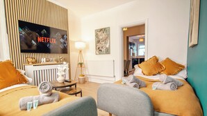 Et hotelværelse med et fjernsyn der viser Netflix-logoet, en seng med et gult tæppe, en grå lænestol og et lille bord med en lampe og pynt.