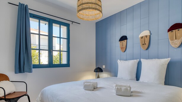 Een slaapkamer met een groot bed, een stoel, een raam met gordijnen, een lamp en maskers aan de muur.