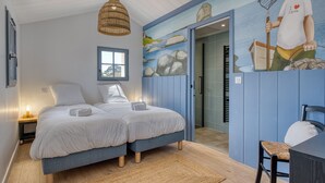 A bedroom with two beds, a woven pendant light, a painting of a fisherman, and a view into the bathroom. - Bains de Mer House & Jacuzzi steps from the beach, Walk to town, Sleeps 6 (Saint-Gilles-Croix-de-Vie)