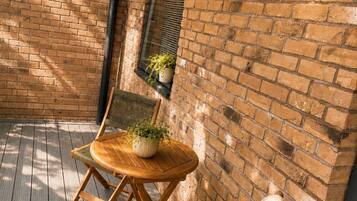 Une terrasse en bois avec deux chaises et une table, un mur en briques et une fenêtre avec des stores.