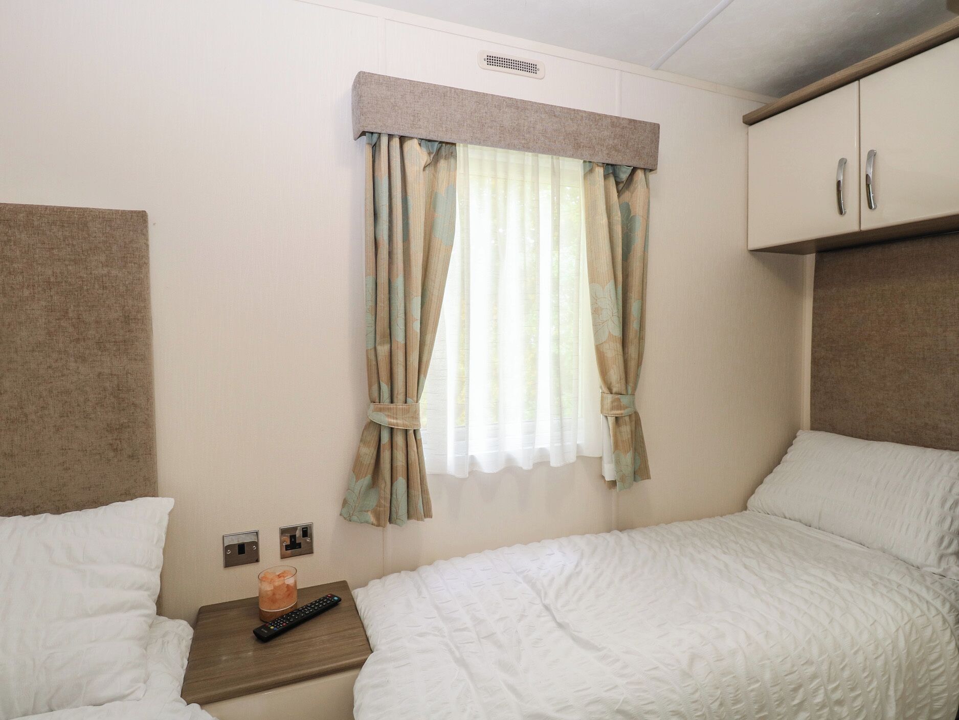 Ein Hotelzimmer mit zwei Betten, einem Nachttisch mit Fernbedienung und einem Fenster mit Vorhängen.