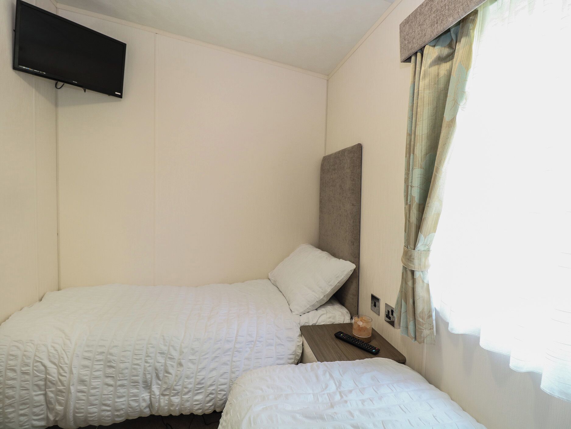 A small, single-room hotel room with two beds, a television mounted on the wall, a bedside table with a remote control, and a window with curtains.