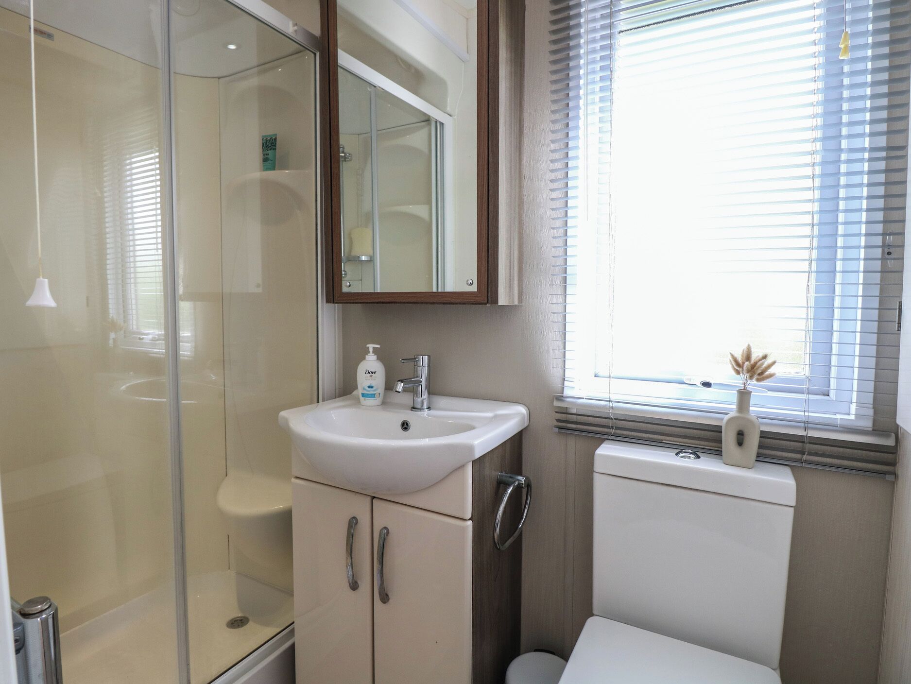 A bathroom with a glass shower enclosure, a sink with a mirror above it, and a window with blinds.
