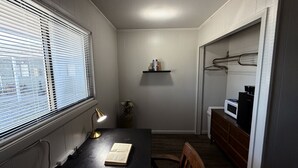 Een kleine kamer met een bureau, een lamp, een stoel, een klein schap met boeken, en een magnetron op een dressoir.