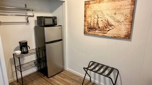 A compact kitchenette with a microwave, coffee maker, and refrigerator. A metal shelf unit is against the wall, and a bench is placed near the door. A vintage map artwork is on the wall. - The Captain`s Room at The Cedar (Sandusky)