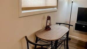 A room with a round wooden table, two chairs, a window with blinds, a floor lamp, and a TV stand. - The Captain`s Room at The Cedar (Sandusky)