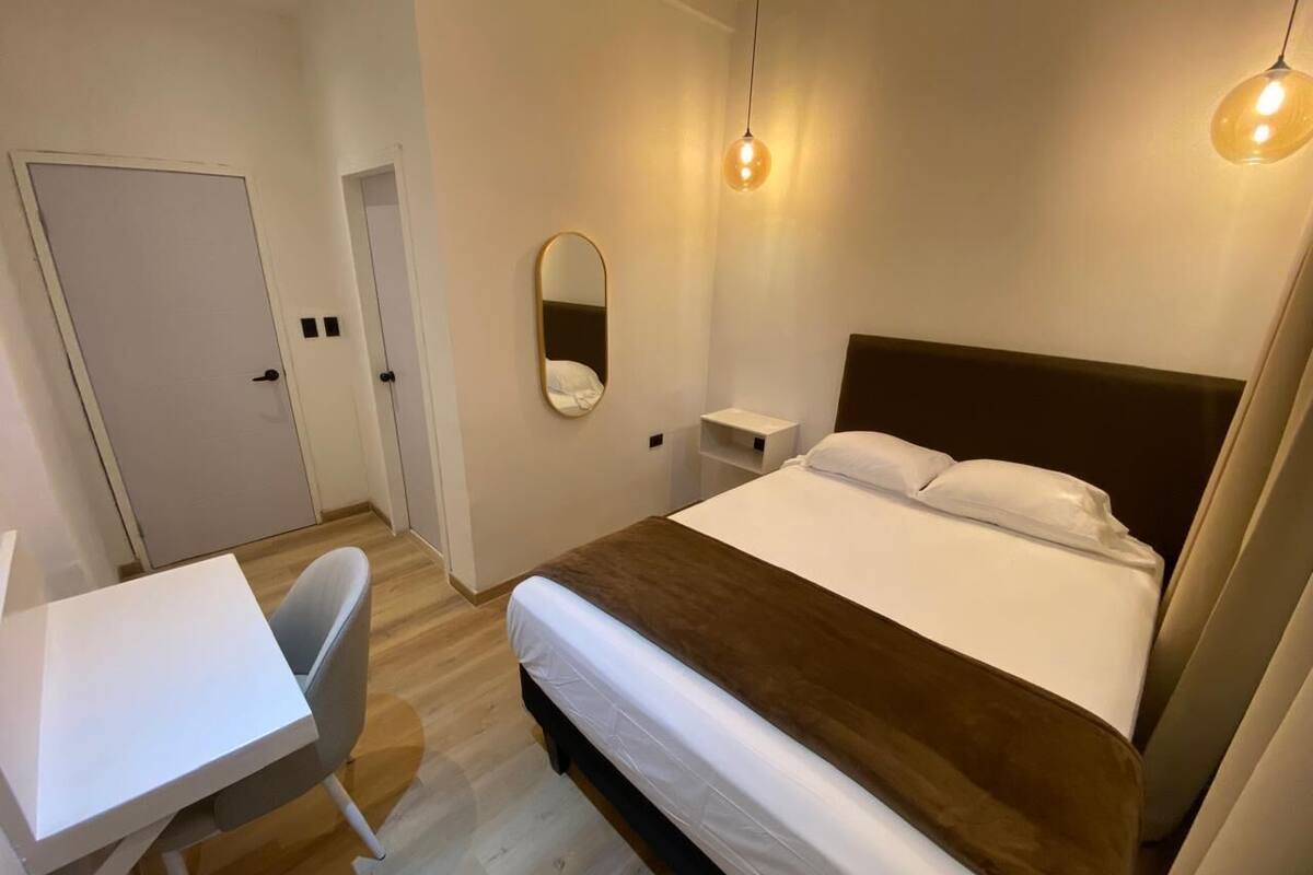 A hotel room with a bed, bedside table, chair, and a desk.