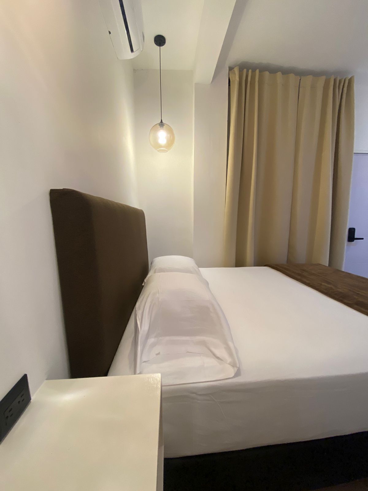 Ett hotellrum med en säng, ett sängbord, en hängande lampa och ett fönster med gardiner.