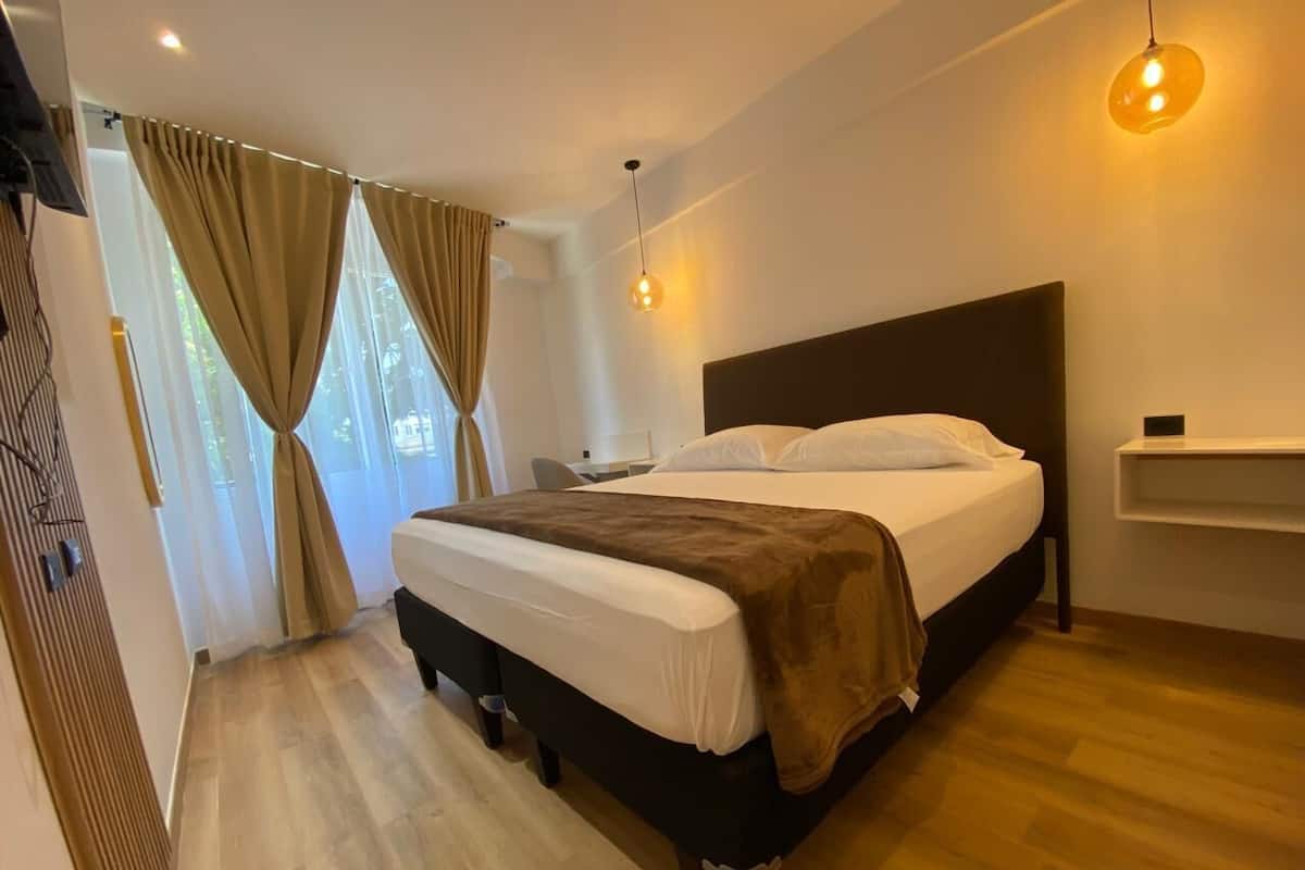 A hotel room with a bed, a desk, a chair, a hanging light fixture, and a window with curtains.