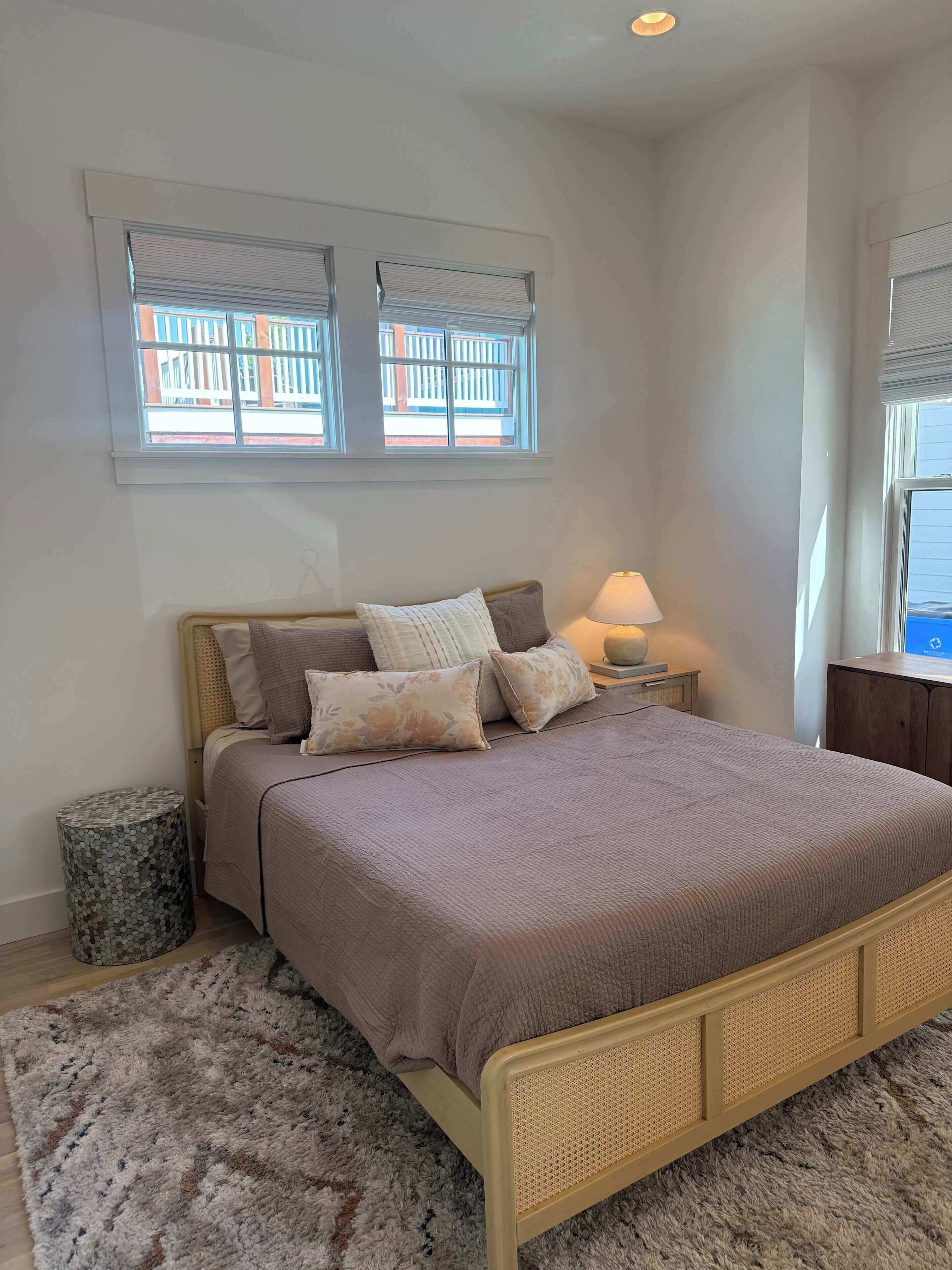 A bedroom with a bed, two windows with blinds, a bedside lamp, and a small round trash bin.