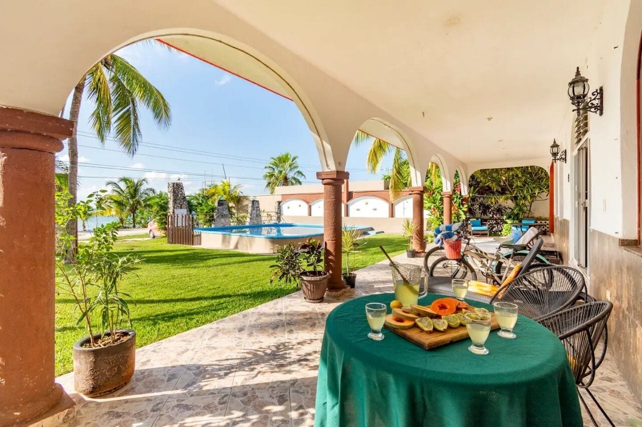A covered outdoor area with a table set for dining, potted plants, and a view of a swimming pool and palm trees.