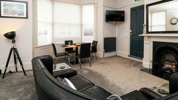 A room with a fireplace, a TV mounted on the wall, a dining table with chairs, and a black leather sofa. - Craven Suite (Scarborough)