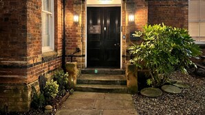 A brick building with a black door, a sign above the door, and a small garden area with a tree and stepping stones. - Craven Suite (Scarborough)