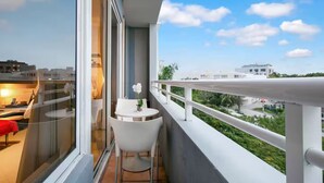 Un balcon avec une chaise blanche et une petite table sur laquelle est posé un vase de fleurs, donnant sur un paysage urbain avec des bâtiments et de la verdure.