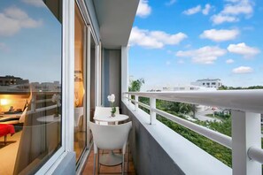 Ein Balkon mit weißem Stuhl und kleinem Tisch, auf dem eine Blumenvase steht, mit Blick auf eine Stadtlandschaft aus Gebäuden und Grünflächen.