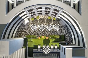 Eine moderne architektonische Struktur mit geschwungenem Design, die einen zentralen Sitzbereich mit einem gemusterten Boden umfasst, umgeben von einer Reihe von Bögen und geometrischen Mustern an den Wänden.