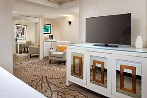 Ein Hotelzimmer mit Glastisch für den Fernseher, einem Flachbildfernseher, einer weißen Kommode mit Holzakzenten, einem gemusterten Teppich und einem Sessel mit gelbem Kissen.