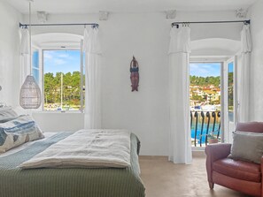 A bedroom with a bed, a chair, a hanging light fixture, a view of a pool and buildings through a window. - Seafront — A Place Like No Other (Stari Grad)