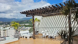 Eine Dachterrasse mit Holzboden, weißen Lounge-Stühlen, einer Pergola und einer Topfpflanze.