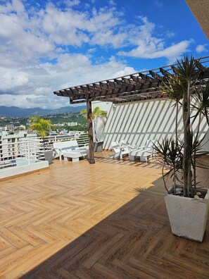 A rooftop terrace with a wooden floor, white lounge chairs, a pergola, and a potted plant. - LUXURY 1 BEDROOM APT  NEW KINGSTON  WITH 24 HRS SECURITY, GYM AND ROOF TOP POOL (Kingston)