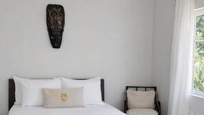 Een bed met wit beddengoed, een houten hoofdbord, een decoratief object aan de muur, een stoel en een raam met gordijnen.