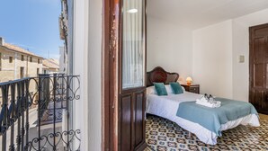 A bedroom with a bed, a nightstand, a window with curtains, a patterned floor, and a balcony with a railing.