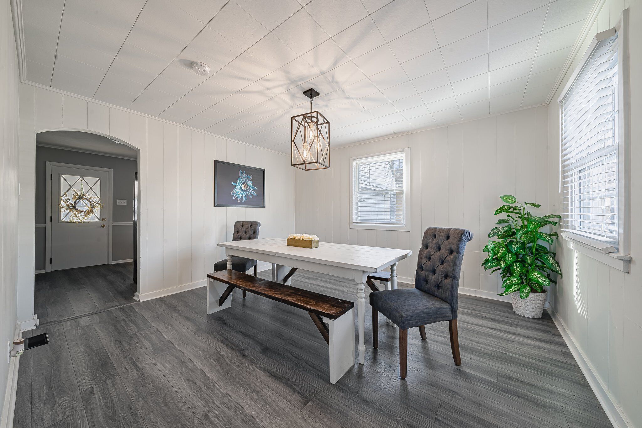 A dining area with a white table, bench, and chair, a hanging light fixture, a potted plant, and a door leading to another room.